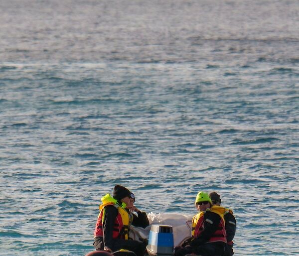 Four personnel in dry suits and life jackets, sitting on sides of IRB, as it travels out to sea away from camera.