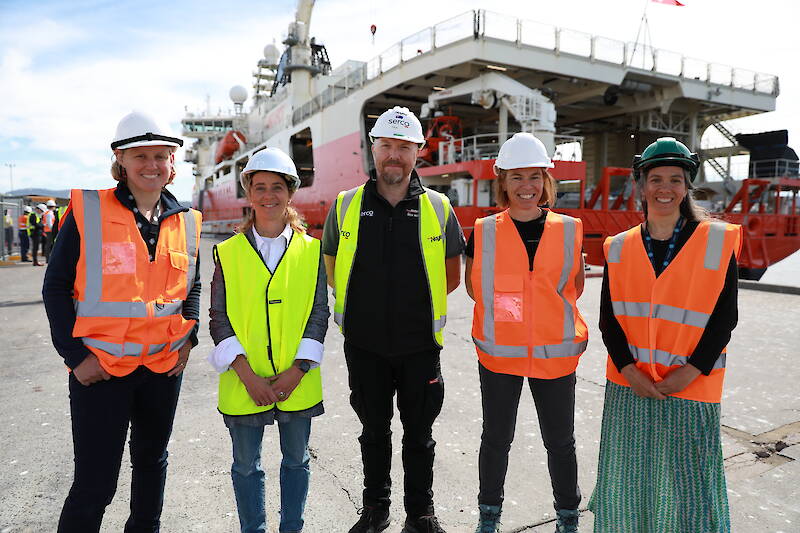 Four women and one man wearing high visibility jackets stand in a line in front of icebreaking ship RSV Nuyina.