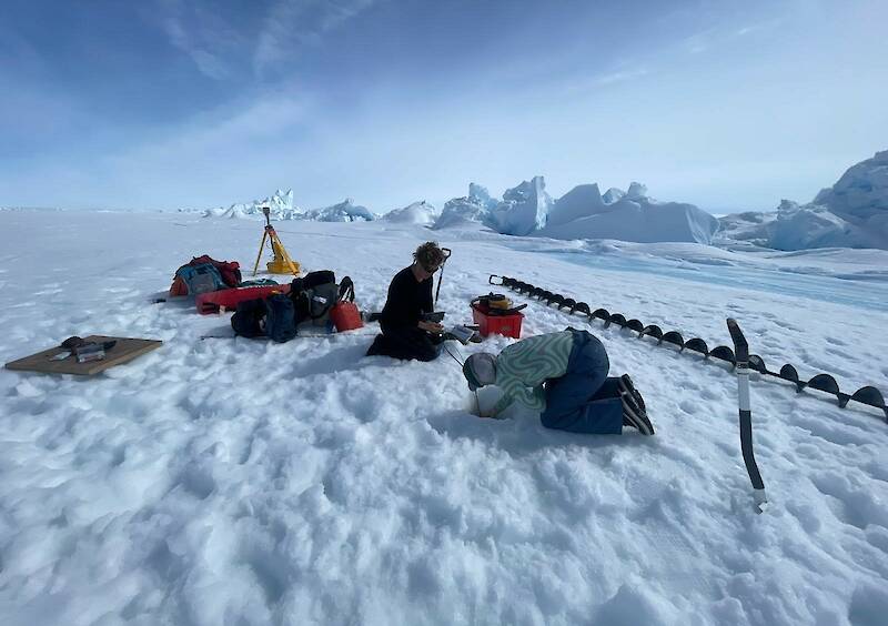 Two people kneeling on a frozen lake. One person is placing a string of sensors in to a hole they have drilled through the ice.