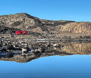 Reflections of a rocky mountain range in a still, blue body of water. A red hut sits on the shore.
