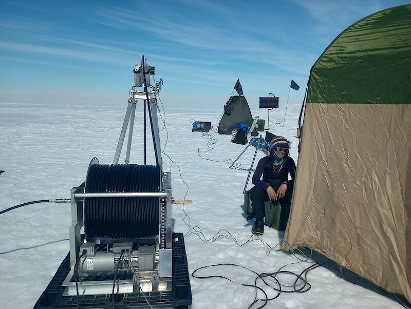 A woman sits on a box beside a tent, monitoring a winch connected to a hot water drill.