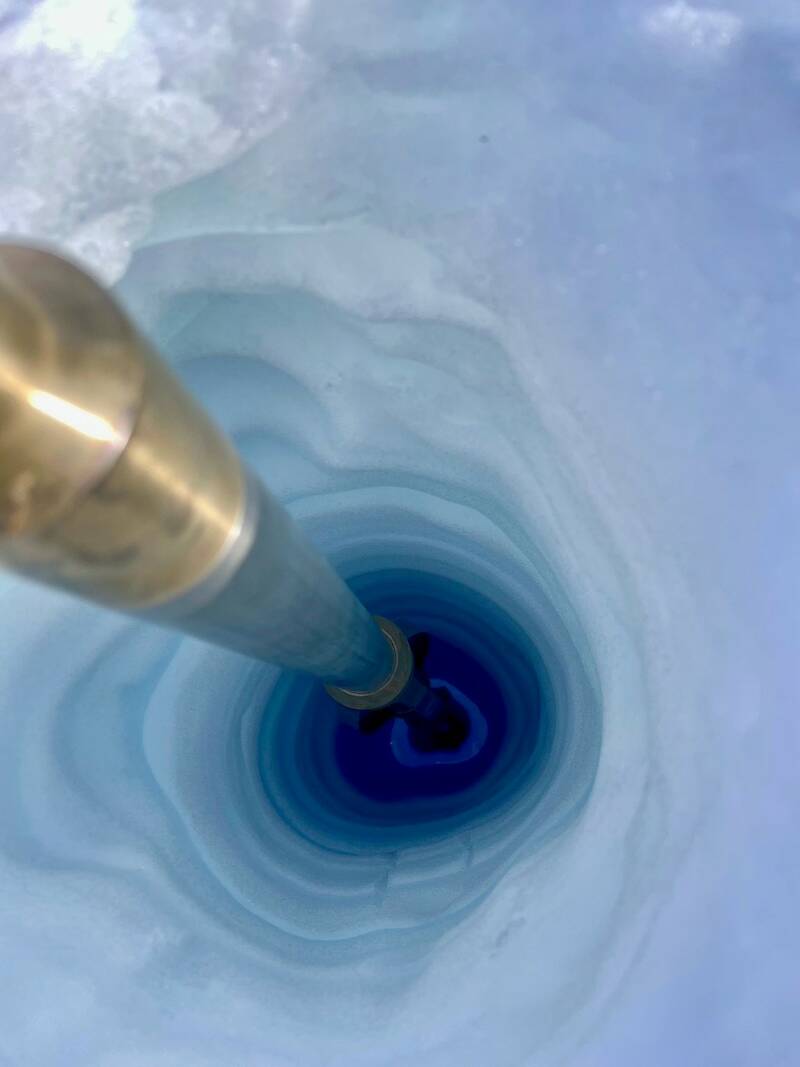 A narrow, deep hole melted in the surface of a glacier. The hole reveals various shades of blue as it gets deeper, from pale near the surface to deep blue towards the base. A metal pipe used to melt the hole is visible