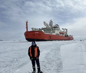 A person in hi vis stands on the ice with a big red ship in the background