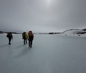 Three people walk across a frozen plain under a faint sun