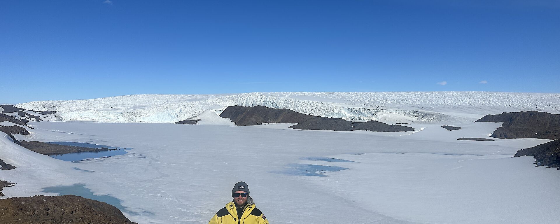 A man in yellow Antarctic clothing stands on a rocky hill with a vista of blue sky and snow behind him