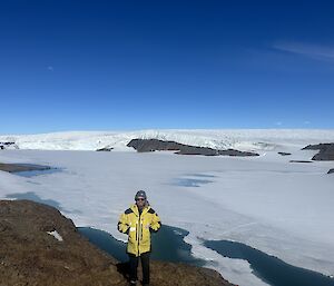 A man in yellow Antarctic clothing stands on a rocky hill with a vista of blue sky and snow behind him