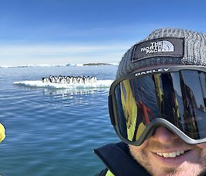 A smiling man with snow goggles on standing in front of open water and an ice floe with penguins on it