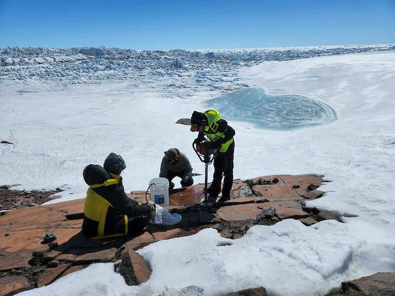 Three people on a small, exposed area of bedrock, surrounded by ice.