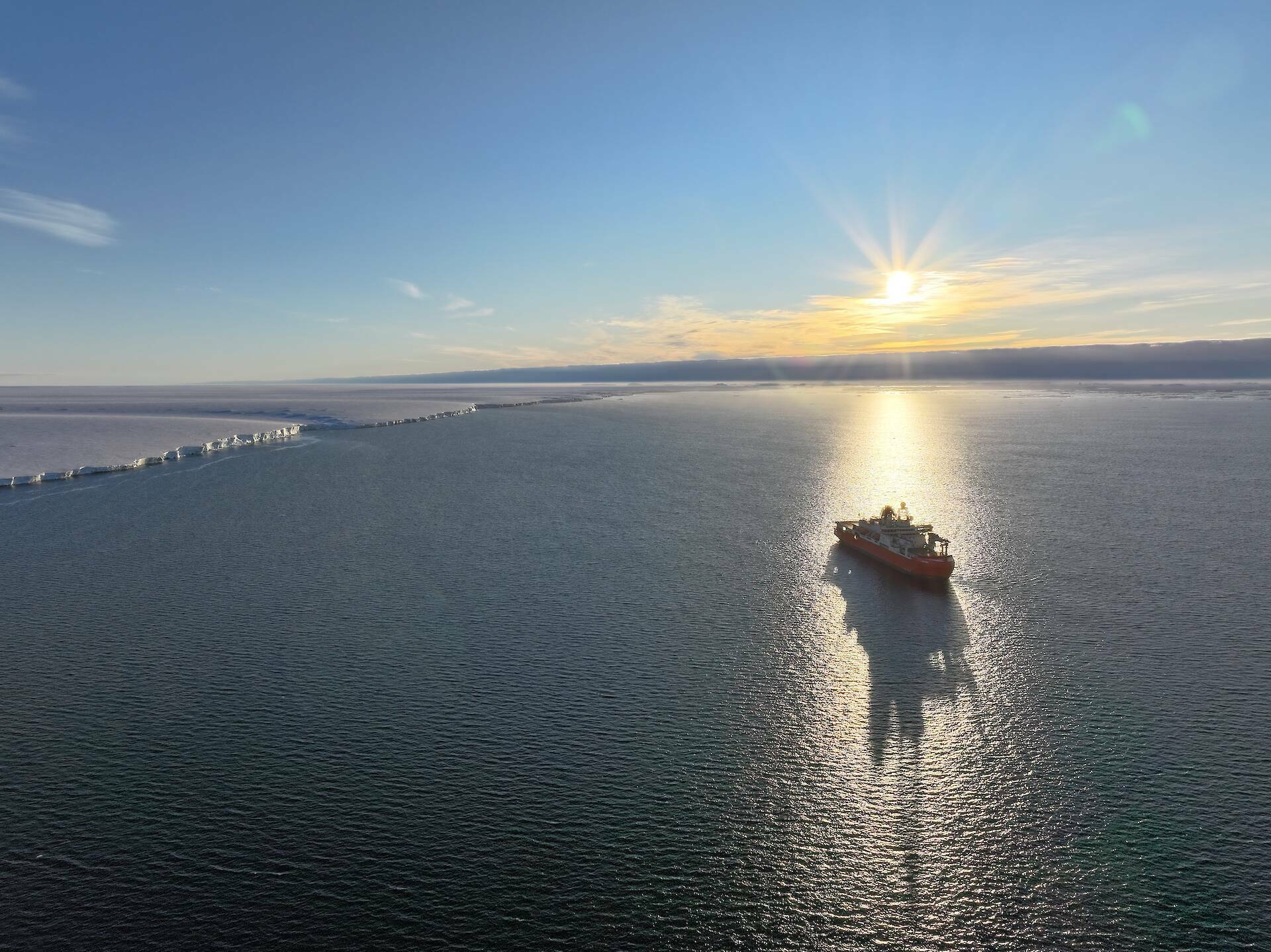 Icebreaking ship RSV Nuyina backlit by the sun, sitting in a calm ocean off the edge of a glacier