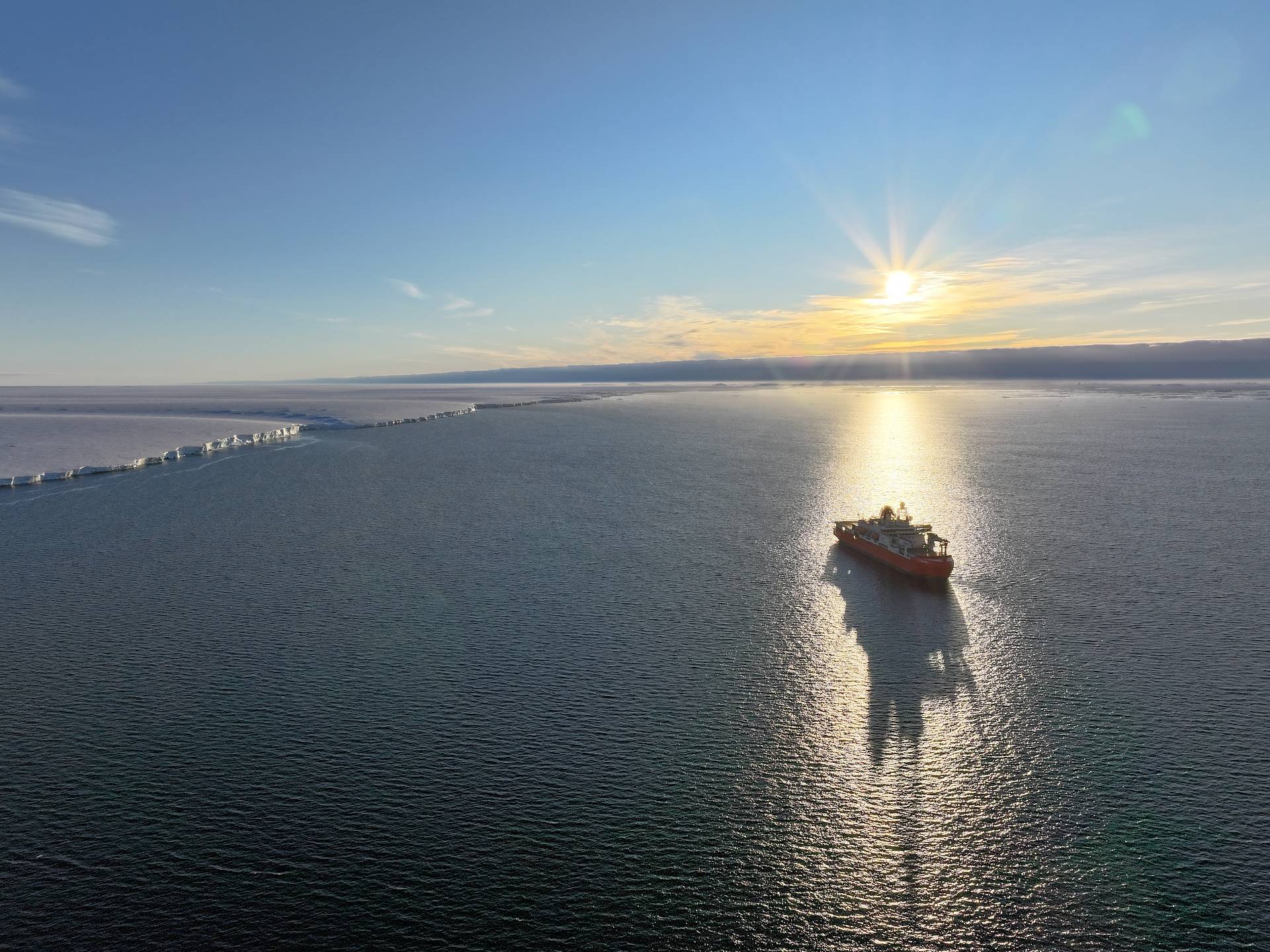 Icebreaking ship RSV Nuyina backlit by the sun, sitting in a calm ocean off the edge of a glacier