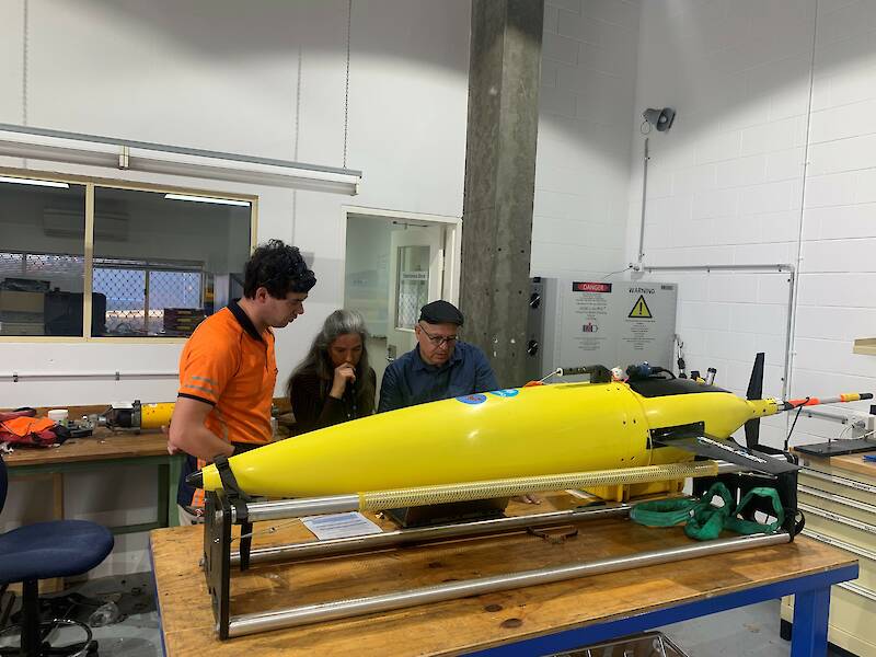 Three people stand beside a bullet-shaped yellow sea glider.