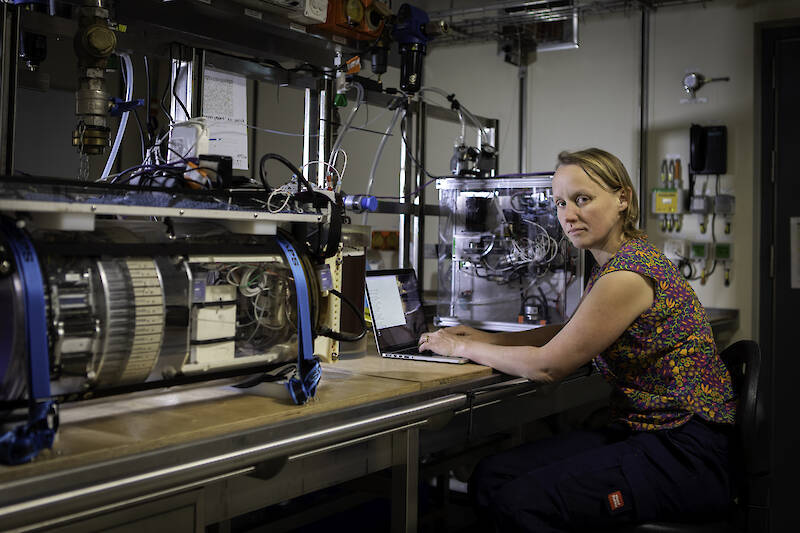 A woman sits with a laptop at a wooden laboratory bench with water sampling equipment cinched tightly to the bench on either side of her.