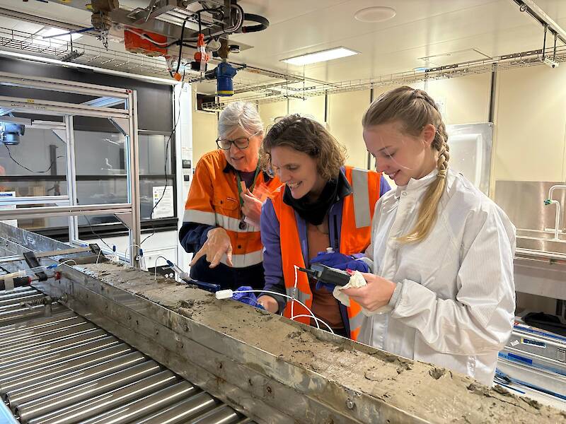 Three women look at a long rectangular length of mud in a metal core barrel.
