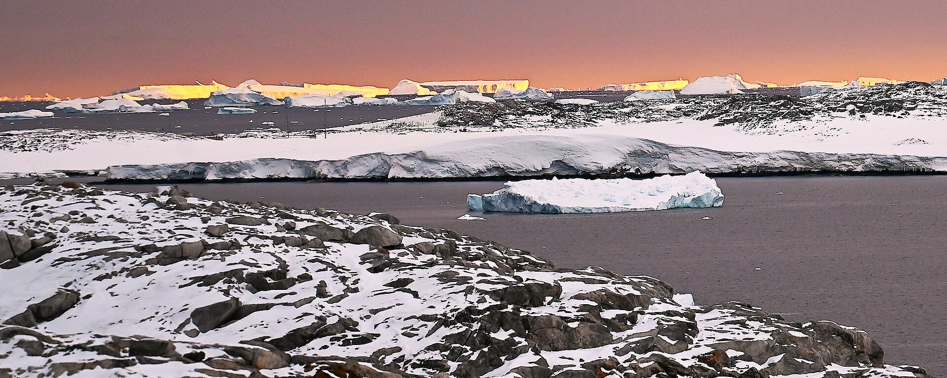 A rocky peninsula part-covered in snow looking out to a sunset on the horizon