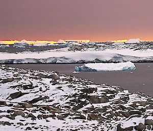 A rocky peninsula part-covered in snow looking out to a sunset on the horizon