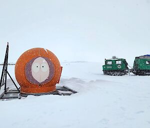 An inflatable 'Kenny' character from the show Southpark poised on the snow with a coupe of snow vehicles in the background