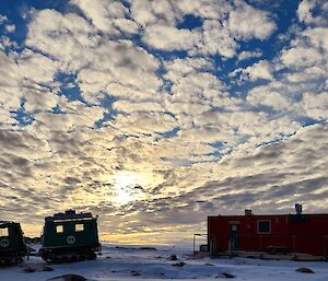 Sunset behind a sky full of small clouds with a small hut and two snow vehicles on the ground