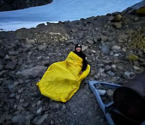 A man in a yellow sleeping bag thing on a rocky slope with ice in the background