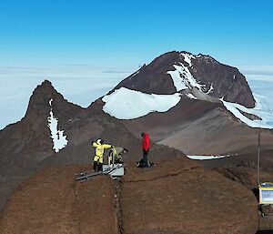 Two people on top of a rock with communications equipment, in front of a grand backdrop of snow-capped mountain peaks