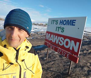 A man in a blue beanie and yellow jacket standing in front of a sign saying 'It's home, it's Mawson'.