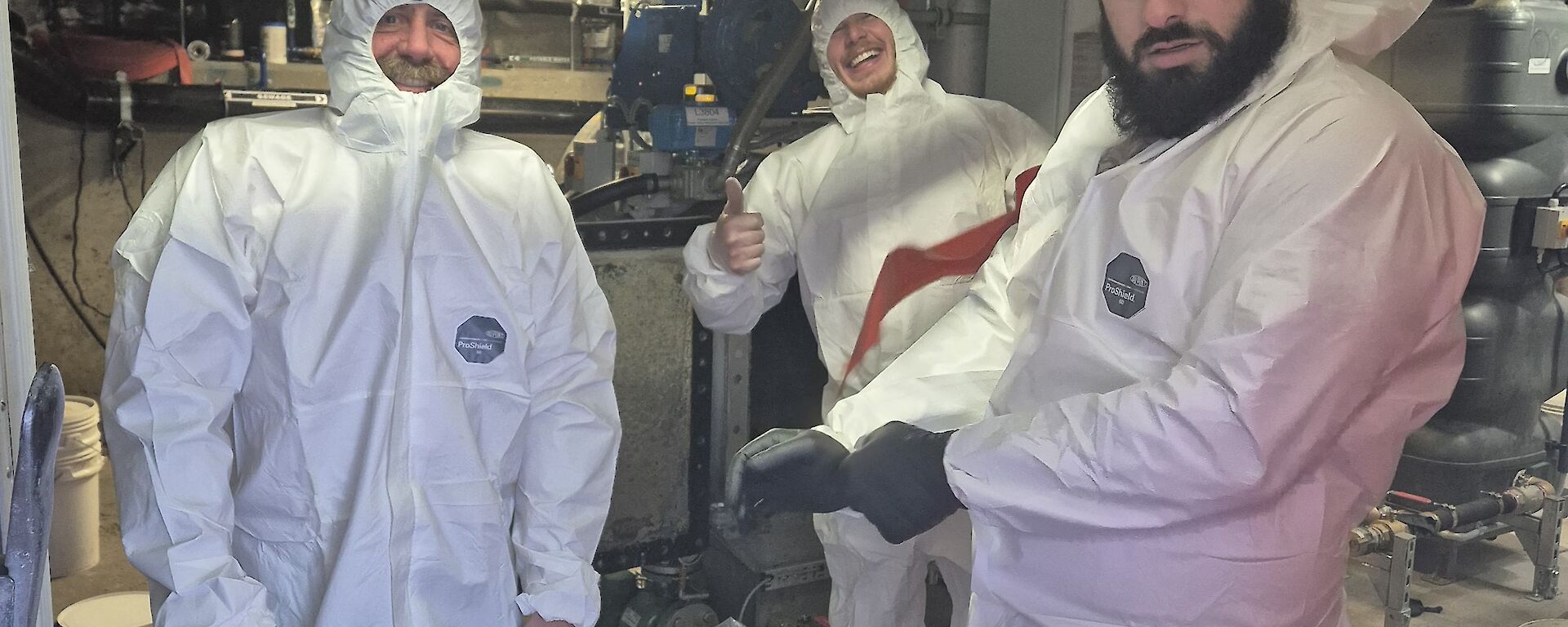 Three expeditioners dressed in white protective suits standing in a workshop cleaning our kitchen grease but still smiling.