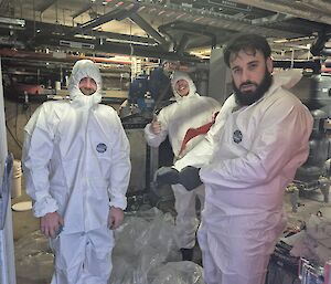 Three expeditioners dressed in white protective suits standing in a workshop cleaning our kitchen grease but still smiling.