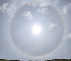 Photo of the sun surrounded by a darker ring, rising over a low-vegetated hillside