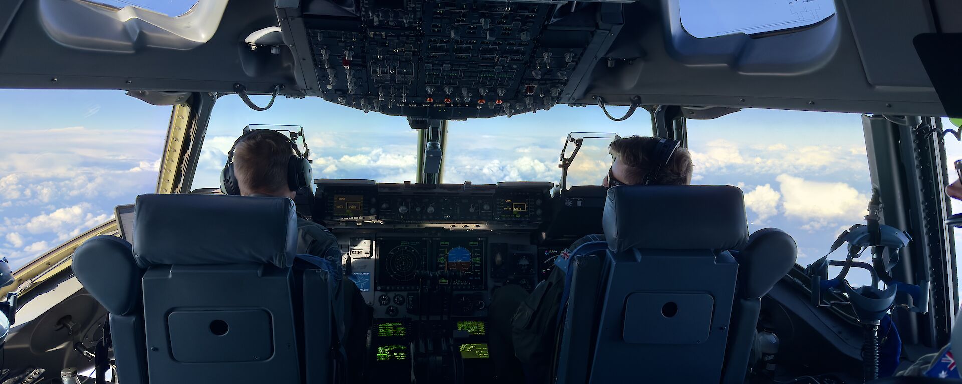 A view from the cockpit of the C17 standing behind the two pilots who are flying the plane and looking out through the windscreen.