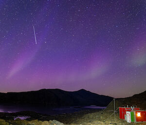 A purple aurora over mountains and a small red hut