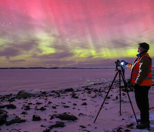 A man standing with a camera on a tripod watching a pink and green aurora