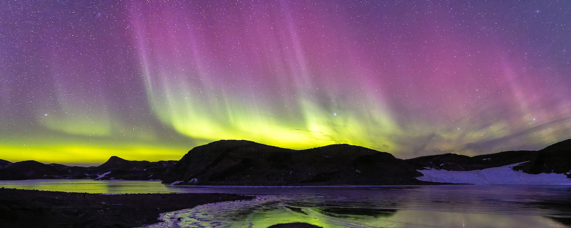 A green and purple aurora makes reflections on a partially frozen lake
