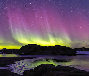 A green and purple aurora makes reflections on a partially frozen lake