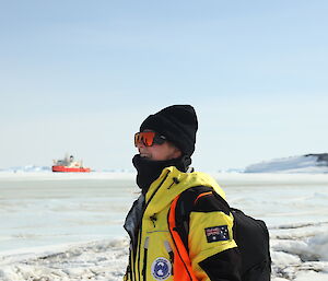 A woman in yellow outdoor gear and a beanie stands on ice with a red ship in the background