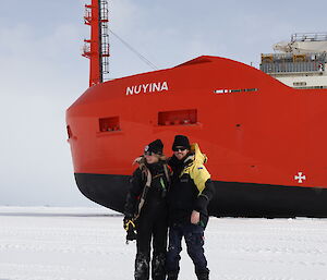 Two people standing on ice in front of a big red icebreaker ship