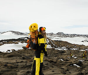 A woman in yellow outdoor gear and snow goggles stands on a rocky slope with snow in the background
