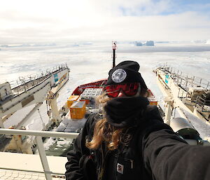 A woman in black takes a selfie while standing on the deck of a ship surrounded by ice and icebergs
