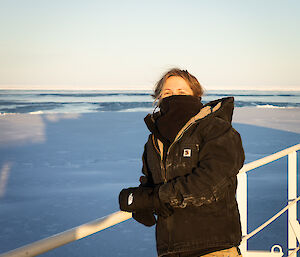 A woman in yellow and black, with a black buff around the face, stands on a ship with blue sea all around