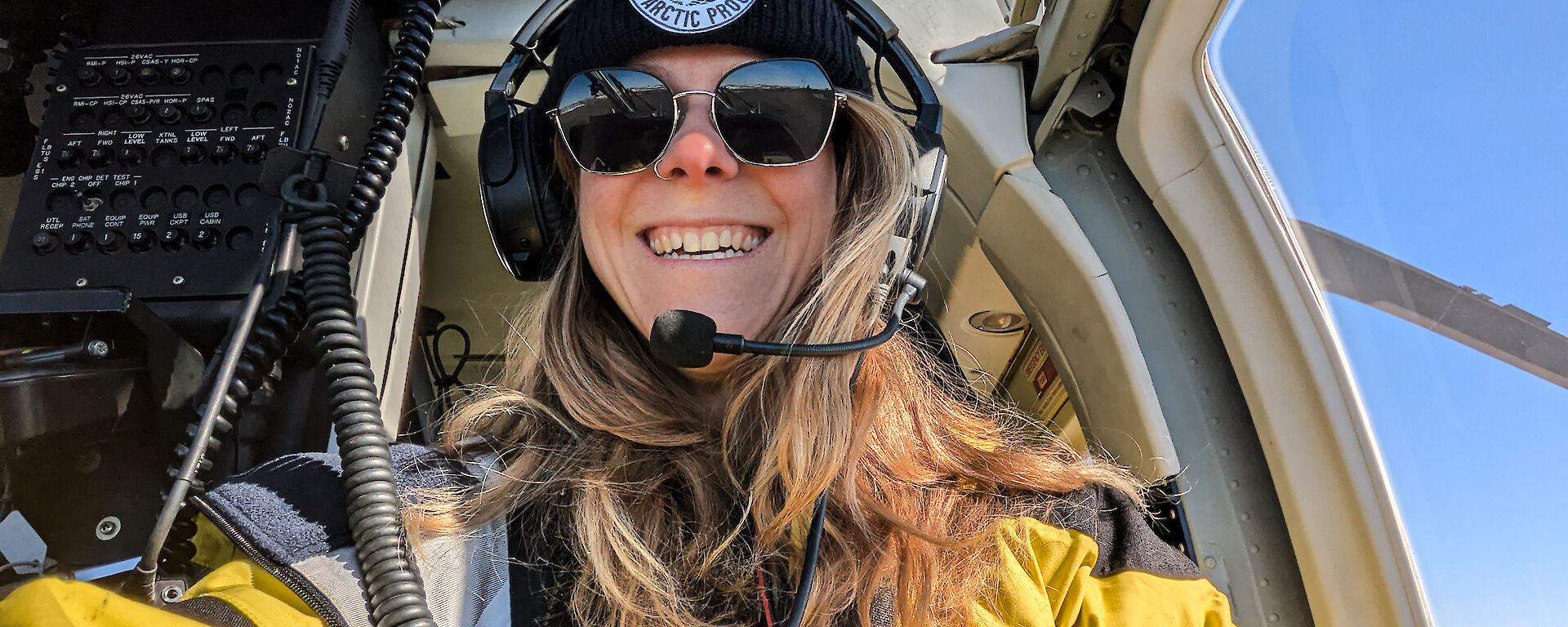 A smiling woman in yellow sitting in a helicopter cockpit with headset, beanie and sunglasses on