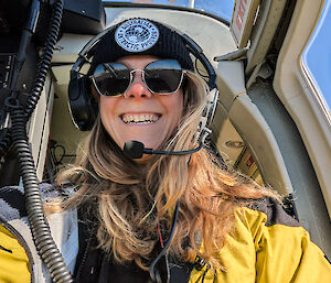 A smiling woman in yellow sitting in a helicopter cockpit with headset, beanie and sunglasses on