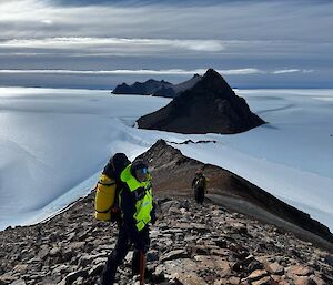 A man in yellow outdoor gear stands on a rocky slope with pyramid-shaped mountains in the background and ice.
