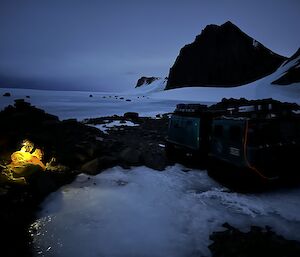A lone yellow light in a tent on a snowy slope in the dim light with black mountains all around