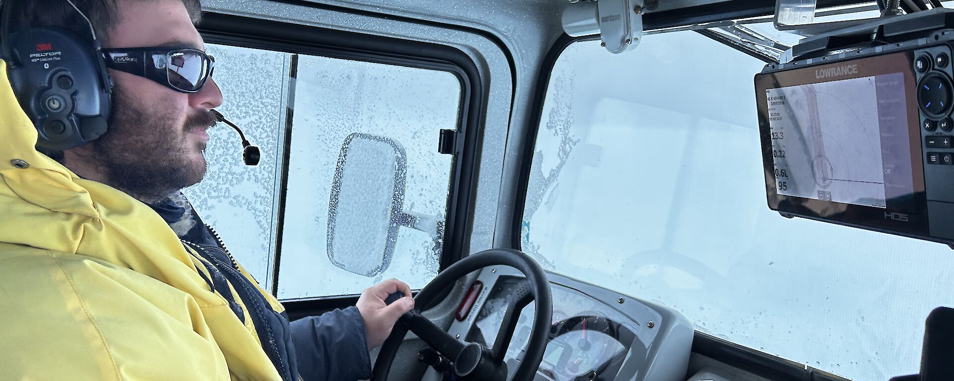 A man in yellow outdoor gear, sunglasses and a headset sitting at the wheel of a truck