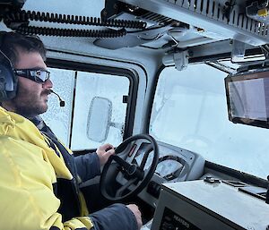 A man in yellow outdoor gear, sunglasses and a headset sitting at the wheel of a truck