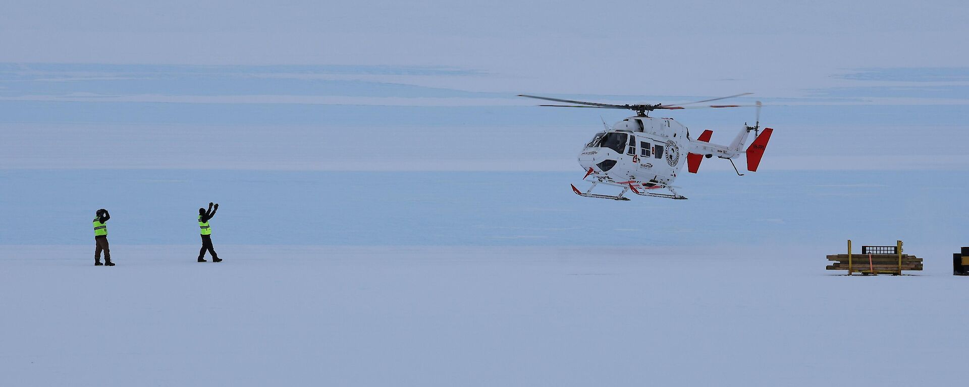 A white and red coloured helicopter hovers above the flat white ice getting waved into land by two expeditioners