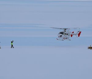 A white and red coloured helicopter hovers above the flat white ice getting waved into land by two expeditioners