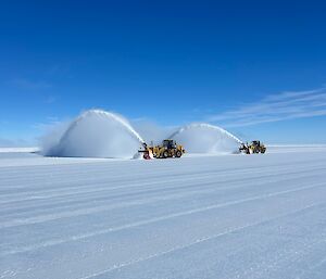 Two large snow grooming machines on the ice runway with visible snow being moved and blown out from the machine