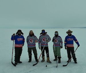 The five Wilkins expeditioners standing on the ice dressed in Casey station ice hockey uniforms holding their ice hockey sticks
