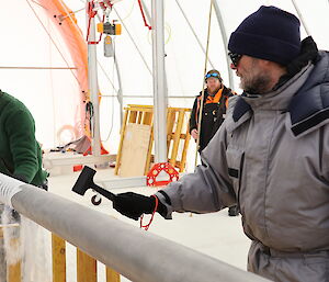 A man removes an ice core from a drill barrel while another man taps the drill barrel with a small rubber hammer