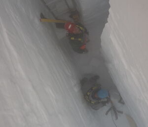 Two men at the bottom of a deep, narrow slot dug out of the ice, moving blocks of ice in to a bag for removal.