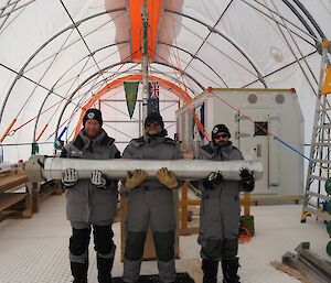 Three men in grey jackets and pants hold a large metal cylinder used to widen a borehole in ice.